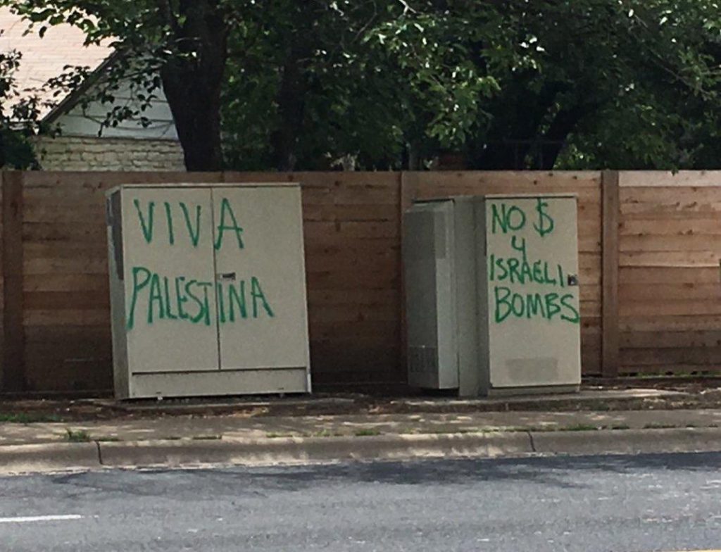 Two grey electrical boxes on the side of a street are decorated with a message in green spray paint. The box on the left reads "Viva Palestina" while the box on the right reads "No $ 4 Israeli Bombs"