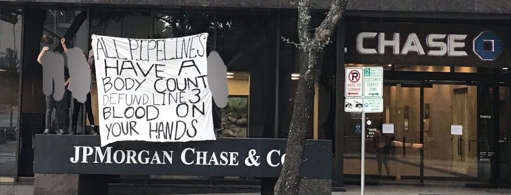 A banner on white cloth is being held in front of a Chase Bank building. The banner is held above a sign reading "JP Morgan Chase & Co," and to the back on the right is an entrance to the building with the Chase Logo. The banner reads: All Pipelines Have a Body Count. Defund Line 3. Blood On Your Hands.