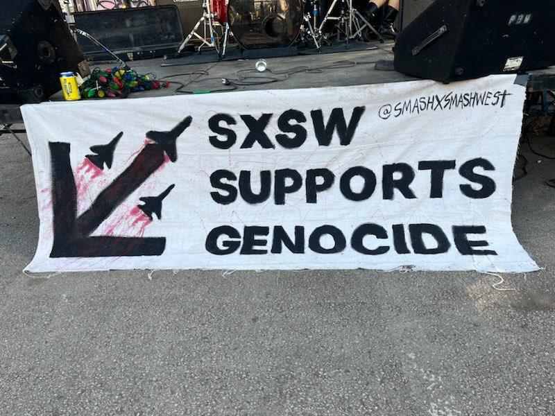 A banner reading "SXSW Supports Genocide" hangs on the front of a music stage. The banner features an arrow pointing down to the left, and 3 fighter planes flying up and to the right with streaks of red blood trailing behind.