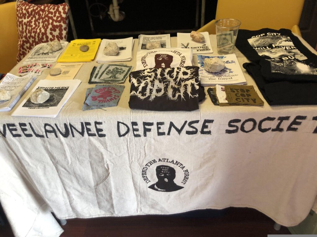 A picture of a table with zines and shirts for sale on top. The table is covered with a banenr cloth that reads "Weelaunee Defense Society." At the center is a picture of someone in a balaclava with the words "Defend the Atlanta Forest" circled around them.